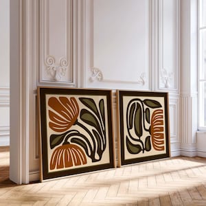 May include: Two framed art prints with abstract floral designs. The artwork features brown, olive green, and cream colours. The frames are a dark brown, and the prints are leaning against a white wall on a wooden floor.