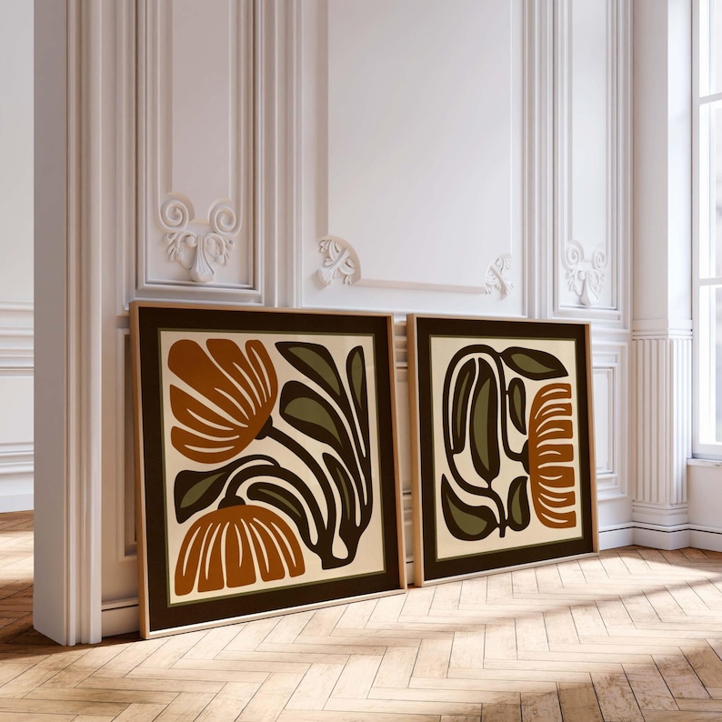 May include: Two framed art prints with abstract floral designs. The artwork features brown, olive green, and cream colours. The frames are a dark brown, and the prints are leaning against a white wall on a wooden floor.