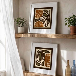 May include: Two framed floral art prints with white borders and brown and olive green designs. The prints are displayed on a rustic wooden shelf, alongside potted plants and decorative bottles. The artwork features abstract botanical illustrations.
