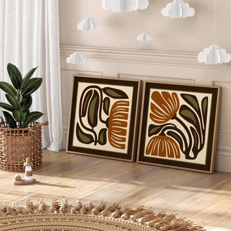 May include: Two framed art prints featuring abstract floral designs in brown, olive green, and cream. The prints are displayed on a wooden floor, with a woven basket and a plant in the background. Paper clouds hang from the ceiling.