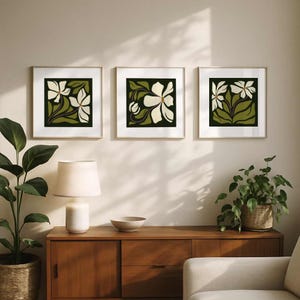 May include: Three framed floral art prints. Each square print features a white flower design with green leaves against a dark green background. The frames have a gold finish. The prints are displayed above a wooden cabinet.