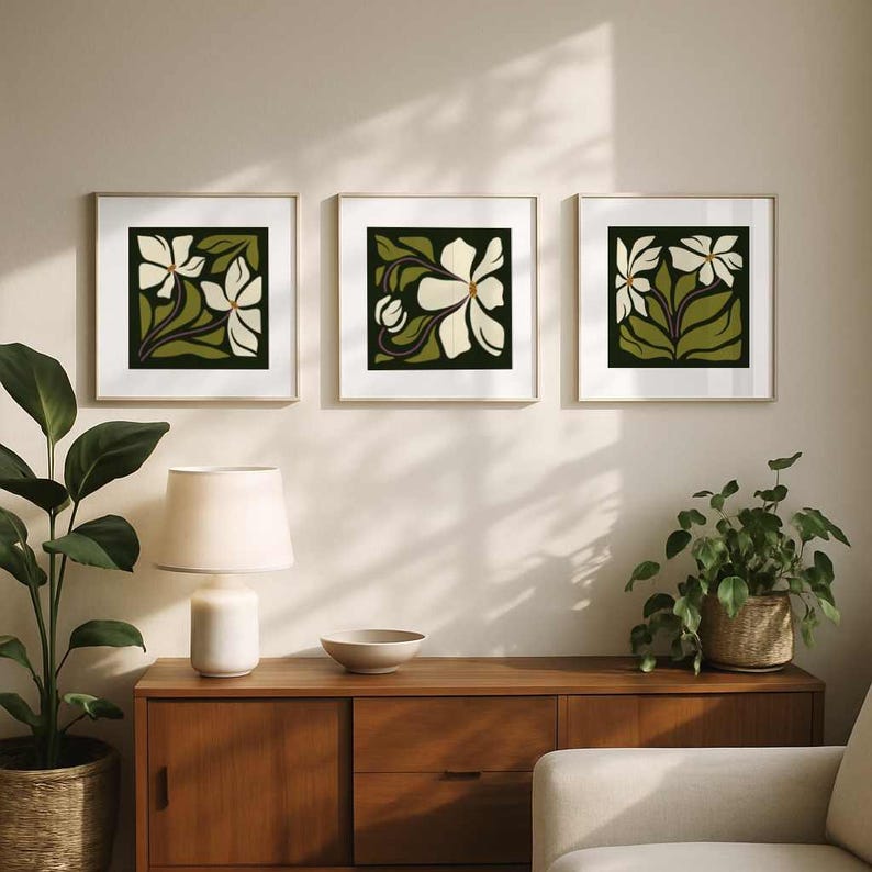 May include: Three framed floral art prints. Each square print features a white flower design with green leaves against a dark green background. The frames have a gold finish. The prints are displayed above a wooden cabinet.