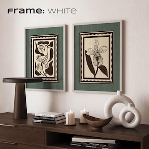 Op de afbeelding: Twee ingelijste botanische kunstprints met witte lijsten en groene passe-partouts. De prints tonen zwarte en cr&egrave;mekleurige bloemmotieven. De tekst "Frame: WHITE" staat bovenaan. Een donkerbruine tafellamp, boeken, kaarsen en decoratieve vazen maken de sc&egrave;ne compleet.
