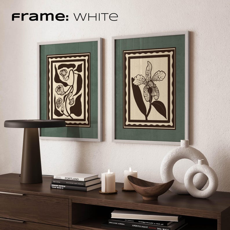 Op de afbeelding: Twee ingelijste botanische kunstprints met witte lijsten en groene passe-partouts. De prints tonen zwarte en cr&egrave;mekleurige bloemmotieven. De tekst "Frame: WHITE" staat bovenaan. Een donkerbruine tafellamp, boeken, kaarsen en decoratieve vazen maken de sc&egrave;ne compleet.