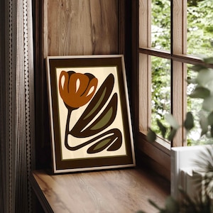 K&ouml;nnte beinhalten: Gerahmter Kunstdruck mit einem stilisierten Blumendesign. Das Kunstwerk zeigt eine braune Tulpe mit gr&uuml;nen Bl&auml;ttern auf cremefarbenem Hintergrund, gerahmt in einem dunkelbraunen Rahmen. Der Druck wird auf einer Holzoberfl&auml;che in der N&auml;he eines Fensters angezeigt.