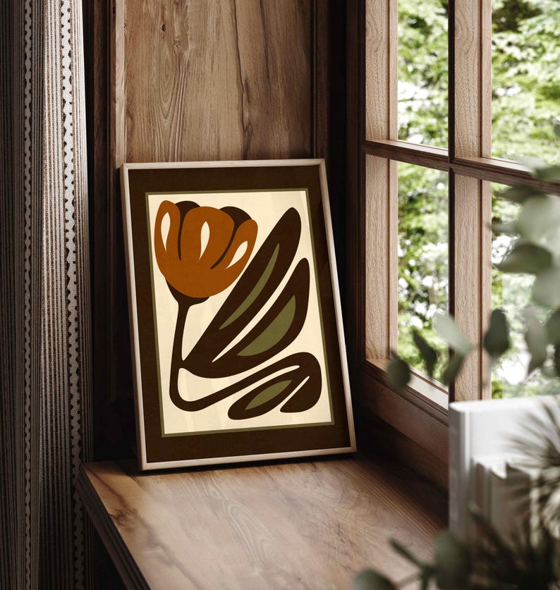 K&ouml;nnte beinhalten: Gerahmter Kunstdruck mit einem stilisierten Blumendesign. Das Kunstwerk zeigt eine braune Tulpe mit gr&uuml;nen Bl&auml;ttern auf cremefarbenem Hintergrund, gerahmt in einem dunkelbraunen Rahmen. Der Druck wird auf einer Holzoberfl&auml;che in der N&auml;he eines Fensters angezeigt.