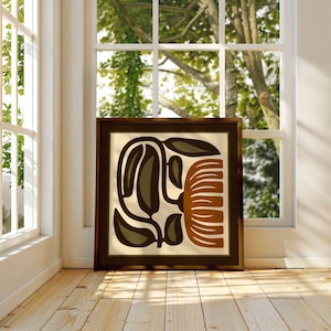 May include: A framed abstract art print featuring a botanical design in shades of brown, olive green, and cream. The artwork is displayed in a room with natural light, near a window with a view of trees.
