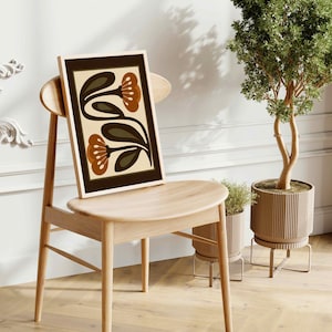 Puede incluir: Una impresi&oacute;n art&iacute;stica enmarcada con un dise&ntilde;o floral retro. La obra presenta flores y hojas marrones y verde oliva sobre un fondo crema. La impresi&oacute;n se muestra en una silla de madera clara. Una planta en maceta est&aacute; en el fondo.