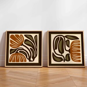 May include: Two framed floral art prints. Each print features a stylized floral design in shades of brown, olive green, and cream. The frames are a light wood colour with a dark brown border.