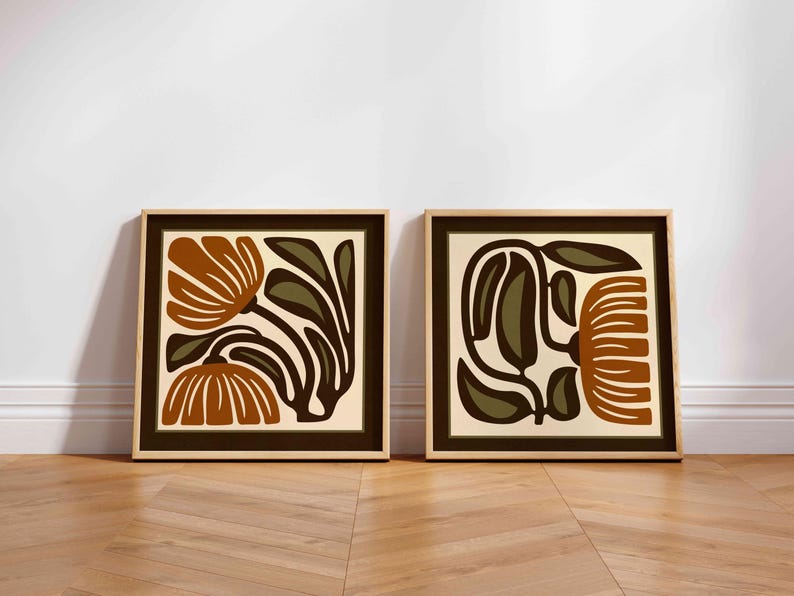 May include: Two framed floral art prints. Each print features a stylized floral design in shades of brown, olive green, and cream. The frames are a light wood colour with a dark brown border.