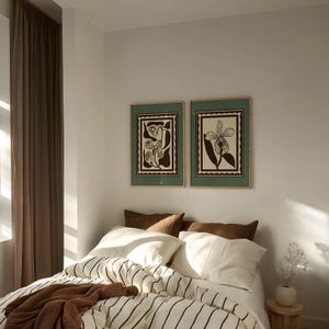 Op de afbeelding: Een slaapkamer met een bed met witte lakens en zwarte strepen, bruine kussens en een bruine deken. Boven het bed hangen twee ingelijste botanische prenten. Links is een bruin gordijn.