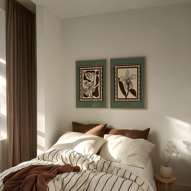 Op de afbeelding: Een slaapkamer met een bed met witte lakens en zwarte strepen, bruine kussens en een bruine deken. Boven het bed hangen twee ingelijste botanische prenten. Links is een bruin gordijn.