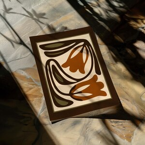 May include: A rectangular art print featuring a stylized floral design. The artwork showcases two brown tulip-like shapes with dark brown and olive green accents against a cream background, framed by a dark brown border. The print is displayed on a textured surface.