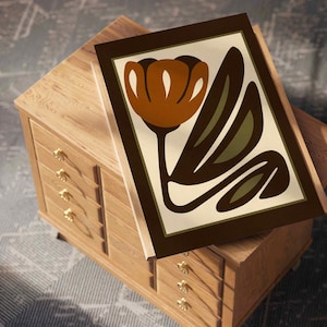 K&ouml;nnte beinhalten: Ein gerahmter Kunstdruck mit einer stilisierten orangefarbenen Tulpe mit gr&uuml;nen Bl&auml;ttern auf cremefarbenem Hintergrund. Der Druck ist in einem dunkelbraunen Rahmen gefasst und liegt auf einer hellen Holzkommode mit goldfarbenen Kn&ouml;pfen. Die Kommode hat mehrere Schubladen.