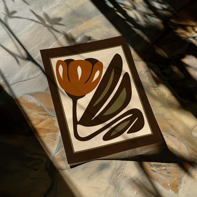 K&ouml;nnte beinhalten: Ein gerahmter Kunstdruck mit einem stilisierten Blumendesign. Das Kunstwerk zeigt eine braune Blume mit olivgr&uuml;nen Bl&auml;ttern auf cremefarbenem Hintergrund, gerahmt in Dunkelbraun. Der Druck liegt auf einer strukturierten Oberfl&auml;che.