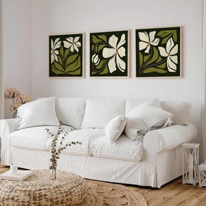 May include: Three framed floral art prints. Each print features a white flower design with green leaves and stems against a dark green background. The frames are a light wood colour. The prints are displayed above a white sofa.