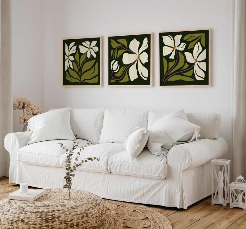 May include: Three framed floral art prints. Each print features a white flower design with green leaves and stems against a dark green background. The frames are a light wood colour. The prints are displayed above a white sofa.