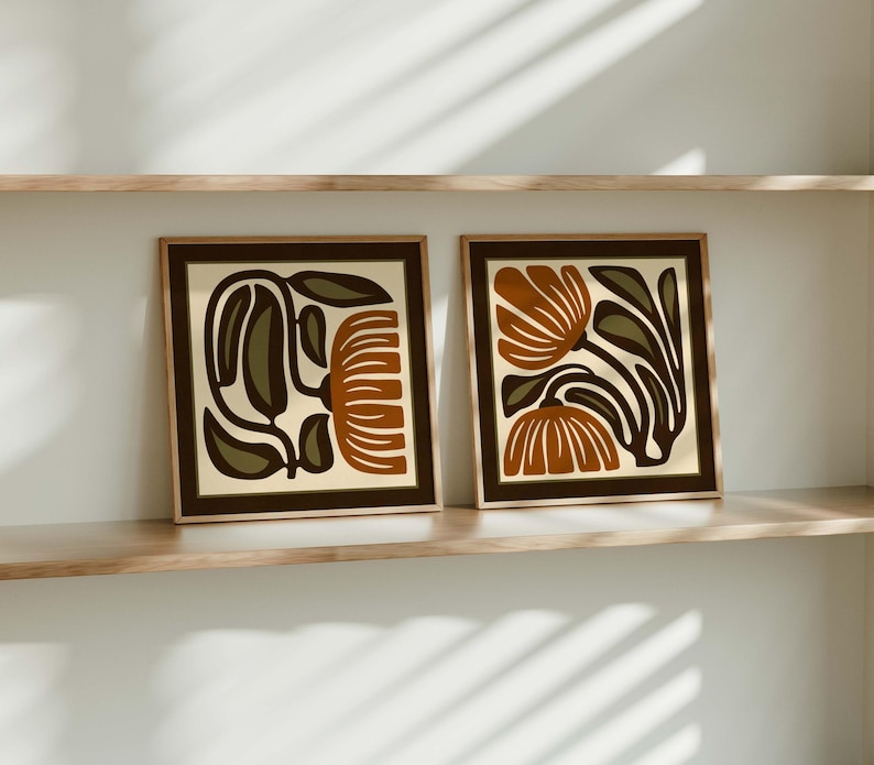 May include: Two framed art prints with abstract floral designs. Each print features a beige background with brown and rust-colored floral shapes and dark green leaves. The frames are brown, and the prints are displayed on a wooden shelf.