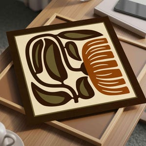 May include: Square framed art print featuring a stylized floral design in shades of brown, olive green, and cream. The artwork is displayed on a light wooden table, with a second frame visible beneath it. A smartphone and book are also on the table.