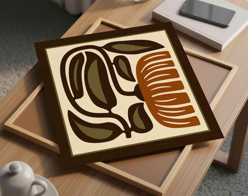 May include: Square framed art print featuring a stylized floral design in shades of brown, olive green, and cream. The artwork is displayed on a light wooden table, with a second frame visible beneath it. A smartphone and book are also on the table.