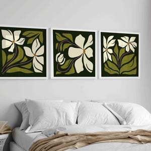 May include: Three framed floral art prints. Each print features a white flower with a yellow centre, green leaves, and a dark green background. The frames are white, displayed above a bed with white linens.