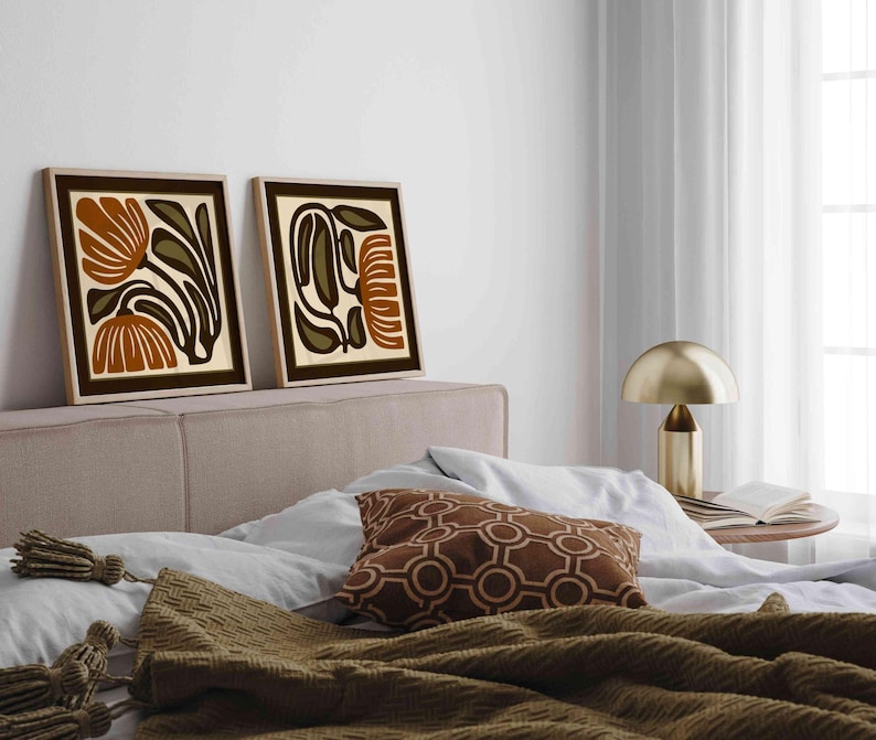 May include: Two framed abstract floral art prints with brown, olive green, and cream colors. The artwork is displayed above a bed with a brown patterned pillow and a gold lamp on a side table.