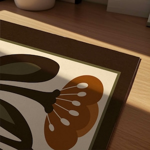 Puede incluir: Primer plano de un estampado floral decorativo sobre una alfombra rectangular. El dise&ntilde;o presenta una flor grande y estilizada en tonos marrones y naranjas sobre un fondo blanquecino, enmarcada por un borde marr&oacute;n oscuro y una fina l&iacute;nea verde.