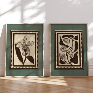 Op de afbeelding: Twee ingelijste botanische kunstprints. Elke print heeft een bloemenontwerp in zwart en beige, tegen een groene achtergrond met een gouden lijst. Het kunstwerk wordt tentoongesteld op een lichte houten vloer tegen een witte muur.