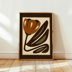 K&ouml;nnte beinhalten: Gerahmter Kunstdruck mit einem stilisierten Blumendesign. Das Kunstwerk zeigt eine gro&szlig;e, rostfarbene Blume mit dunkelbraunen und olivgr&uuml;nen Bl&auml;ttern vor einem cremefarbenen Hintergrund. Der Druck ist in einem Holzrahmen mit dunkelbraunem Rand.