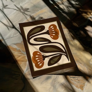 Puede incluir: Una impresi&oacute;n art&iacute;stica rectangular con un dise&ntilde;o floral retro. La obra de arte presenta dos flores estilizadas en tonos marrones y naranjas, con hojas verde oliva, sobre un fondo crema. La impresi&oacute;n est&aacute; enmarcada con un borde marr&oacute;n oscuro.