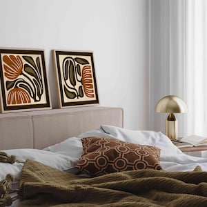 May include: Two framed floral art prints in brown, olive green, and cream tones. The artwork is displayed above a bed with a brown patterned pillow and a gold lamp. The prints have a minimalist, botanical theme.