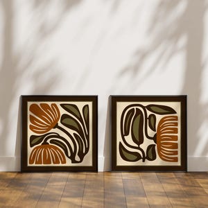May include: Two framed abstract floral art prints. Each print features a brown frame with a cream-colored background and a stylized floral design in shades of brown, olive green, and black. The prints are displayed on a wooden floor against a white wall.