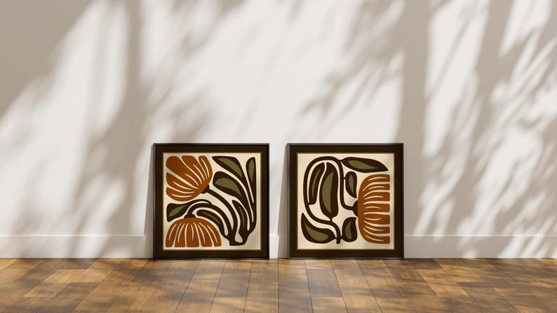 May include: Two framed abstract floral art prints. Each print features a brown frame with a cream-colored background and a stylized floral design in shades of brown, olive green, and black. The prints are displayed on a wooden floor against a white wall.