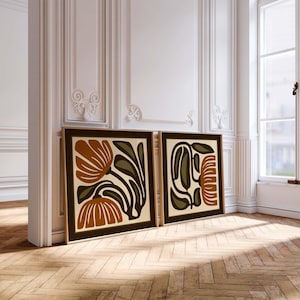 May include: Two framed art prints with floral designs. The artwork features abstract flowers in shades of brown, olive green, and cream, set against a dark brown border. The frames are a light wood color, and the prints are leaning against a white wall.