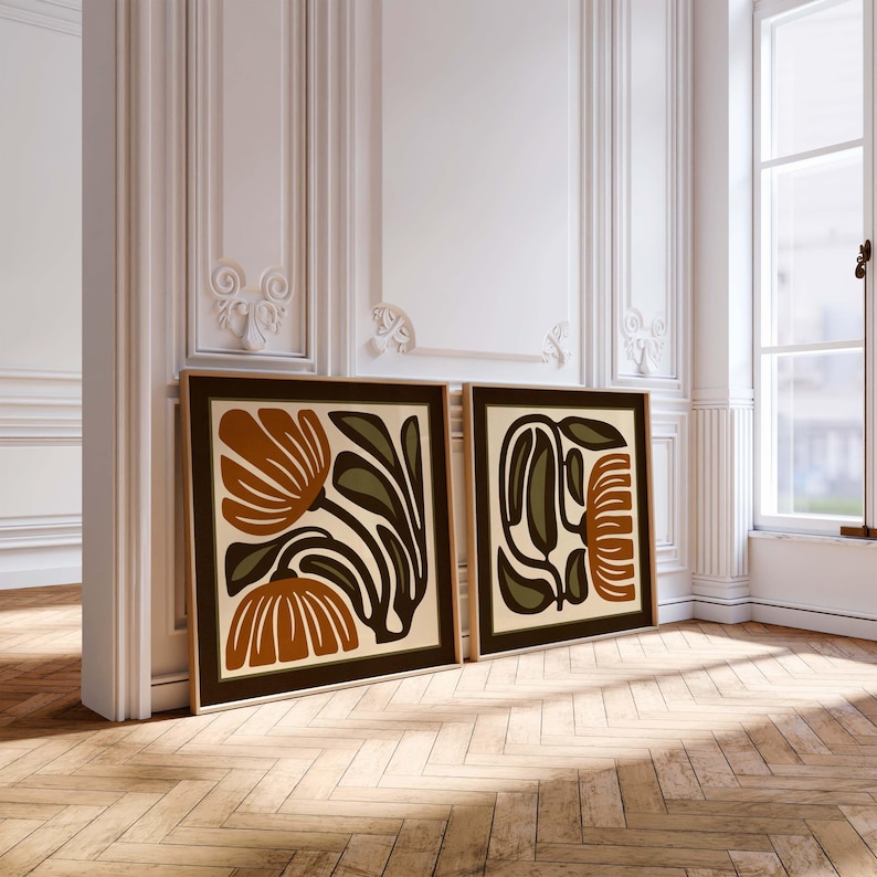 May include: Two framed art prints with floral designs. The artwork features abstract flowers in shades of brown, olive green, and cream, set against a dark brown border. The frames are a light wood color, and the prints are leaning against a white wall.