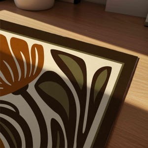 May include: Close-up of a decorative floral print with a retro design. The artwork features a large orange flower and green leaves against a cream background, framed by a dark brown border. The design has a vintage, mid-century modern aesthetic.