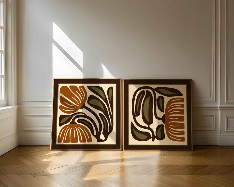 May include: Two framed abstract floral art prints. Each print features a brown frame, a cream-colored background, and stylized flowers in shades of brown, orange, and olive green. The artwork is displayed against a white wall.