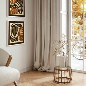 May include: Two framed abstract floral art prints in brown, beige, and olive green hang on a white wall. A beige chair with wooden arms sits in the corner. A small round table with a glass top and a vase with branches is in front of a window.