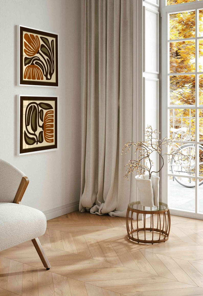 May include: Two framed abstract floral art prints in brown, beige, and olive green hang on a white wall. A beige chair with wooden arms sits in the corner. A small round table with a glass top and a vase with branches is in front of a window.