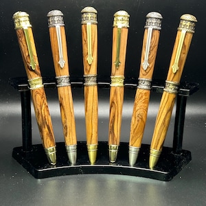 May include: Six handcrafted wooden pens with gold and silver accents are displayed on a black stand. The pens have a natural wood grain pattern, with ornate metal detailing on the caps and tips. The pens are a mix of gold and silver.