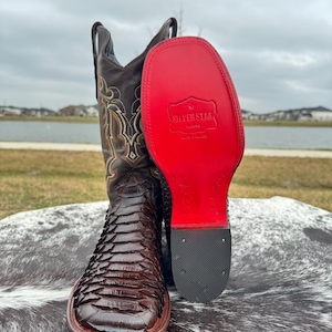 Op de afbeelding: Bruine leren cowboylaarzen met een slangenhuidpatroon en een rode zool. De zool heeft de tekst "Silver Star Boots" erop gedrukt.