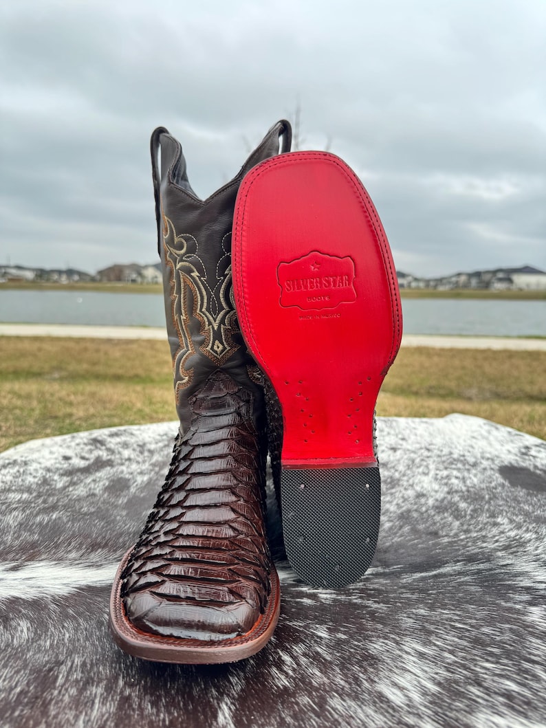 Pu&ograve; includere: Stivali da cowboy in pelle marrone con motivo a pelle di serpente e suola rossa. La suola ha la scritta "Silver Star Boots" stampata.