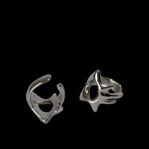 Puede incluir: Dos anillos plateados con diseños abstractos y calados. Uno tiene una forma curva, similar a un ear cuff, mientras que el otro presenta una forma geométrica más estructurada. Ambos anillos están sobre un fondo negro.
