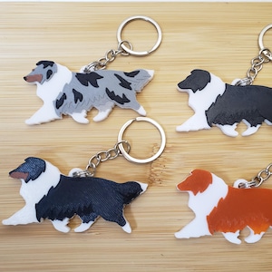 Op de afbeelding: Vier sleutelhangers in de vorm van een hond in verschillende kleuren: blauw merle, zwart en wit, effen zwart en rood en wit. Elke sleutelhanger heeft een zilveren sleutelring en ketting. De sleutelhangers liggen op een lichtbruin houten oppervlak.