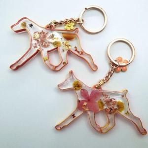 May include: Two dog-shaped keyrings. The keyrings are made of clear resin with dried flowers and gold leaf inclusions. The edges are outlined in a reddish-orange colour, and they have gold-coloured key rings.