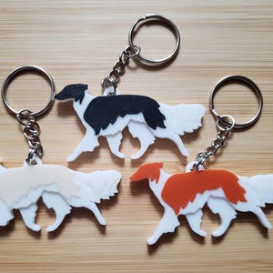 Puede incluir: Tres llaveros con la silueta de un perro Borzoi. Un llavero tiene un perro negro y blanco, otro un perro crema y blanco, y el tercero un perro naranja y blanco. Cada llavero incluye una anilla de metal plateado y una cadena corta.