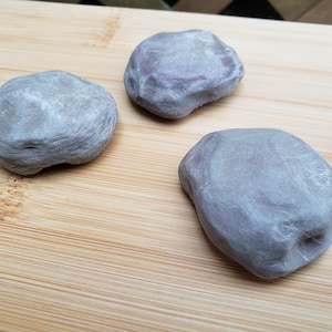 May include: Three smooth, rounded, gray objects with a layered, fibrous texture. Each object has an irregular, flattened shape with visible fine lines across the surface, resembling a dense, molded material or a compressed, textured substance.