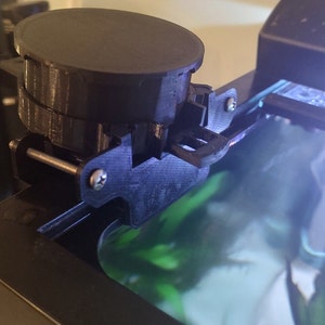 以下が含まれることがあります： 黒い3Dプリントパーツのクローズアップ。ネジと小さな丸い黒いオブジェクトが取り付けられています。パーツは黒い表面に取り付けられています。