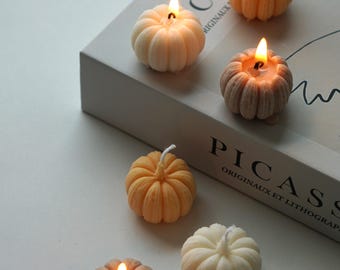 Pode incluir: Seis pequenas velas em forma de abóbora com textura canelada. As velas estão acesas e são de vários tons de laranja, marrom e branco. As velas estão dispostas sobre uma superfície branca com um livro no fundo.