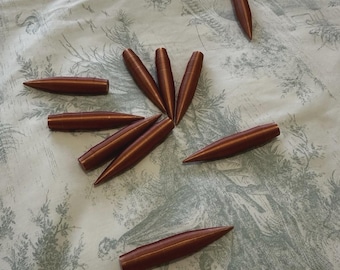 Può includere: Dieci oggetti marroni a forma di proiettile, con punte, sono disposti su un tessuto chiaro a fantasia. Gli oggetti sembrano essere in legno o un materiale simile. Disposizione a cerchio.